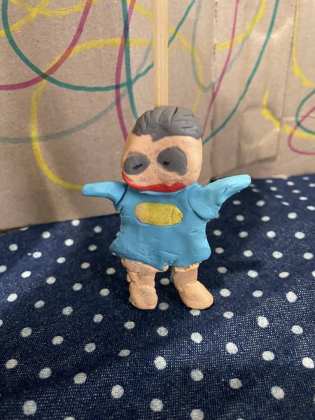 Clay model of a person with grey hair and eyes, wonky smile and blue clothing. Their arms are in the air